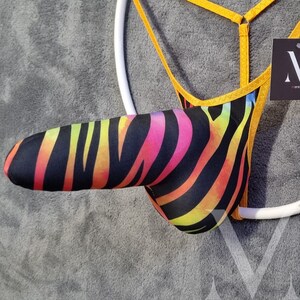 May include: A black and rainbow zebra print jockstrap with yellow straps. The jockstrap is hanging on a white ring.
