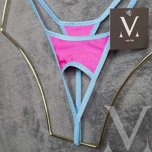 May include: A pink thong with light blue trim and straps. The lingerie is displayed on a gold-colored hanger against a gray background. A tag with a stylized "V" and the text "mens jew" is attached.