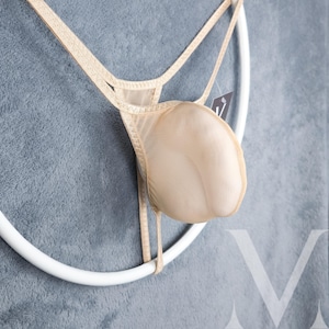 Pode incluir: Um fio dental de malha cor de pele com acabamento dourado e um anel branco preso na parte de trás. O fio dental está pendurado em um anel branco.