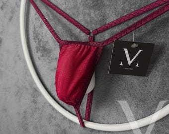 Puede incluir: Un tanga burdeos con tirantes finos y una cintura a juego. La lencería está suspendida de una percha circular blanca. Una etiqueta negra con un logotipo "V" blanco está adjunta.