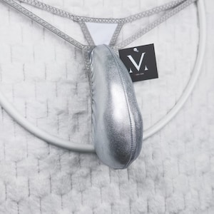 May include: Silver metallic thong underwear with a pouch. The thong has a thin, silver waistband and a white triangle at the front. A black tag with a white "V" logo hangs from the underwear. The background is a white, textured fabric.