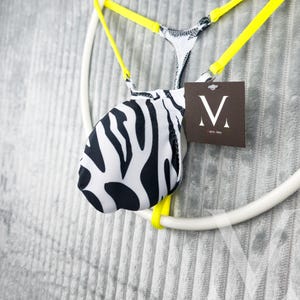 May include: A black and white zebra print thong swimsuit with yellow straps. The swimsuit is hanging on a white circular hanger. A brown tag with a white "V" logo is attached.