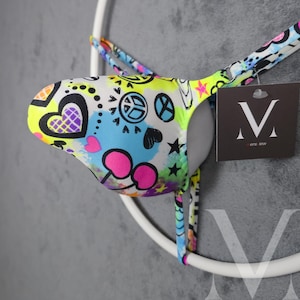 May include: A colorful bikini bottom with a vibrant print featuring hearts, peace signs, and stars. The design incorporates neon yellow, pink, blue, and black elements. The garment has thin straps and a tag with the brand name.