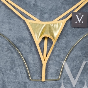 May include: Gold-colored thong underwear with a metallic sheen. The design features a triangular front panel with a keyhole cutout and thin straps. A small tag with a stylized "V" logo is attached.