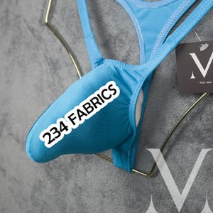 Könnte beinhalten: Ein hellblauer String-Badeanzug mit dem Text "234 FABRICS" auf der Vorderseite. Der Badeanzug wird auf einem goldfarbenen Kleiderbügel vor grauem Hintergrund präsentiert. Ein Etikett mit einem stilisierten "V" ist sichtbar.
