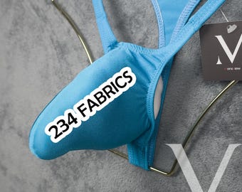 Op de afbeelding: Een lichtblauwe string-badpak met de tekst "234 FABRICS" op de voorkant. Het badpak wordt gepresenteerd op een goudkleurige hanger tegen een grijze achtergrond. Een label met een gestileerde "V" is zichtbaar.