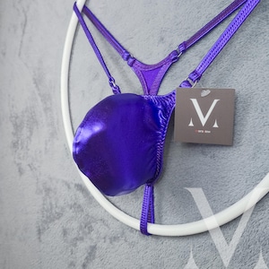 May include: A shiny, deep purple swimsuit bottom with thin straps and a matching top. The garment is hanging on a white circular frame against a gray background. A small tag with a stylized "V" logo is attached.