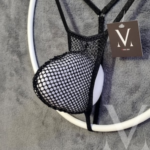 Könnte beinhalten: Schwarze Mesh-Unterwäsche mit weißem Innenfutter und einem Etikett mit der Aufschrift "mers-tow". Die Unterwäsche ist an einem weißen kreisförmigen Rahmen vor grauem Hintergrund aufgehängt. Das Design hat eine netzartige Textur.