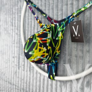 Könnte beinhalten: Ein String-Badeanzug mit einem farbenfrohen Graffiti-Muster in Gelb, Grün und Rot auf dunkelblauem Hintergrund. Die Träger sind mehrfarbig. Ein Etikett mit weißem "V"-Logo hängt am Kleidungsstück.