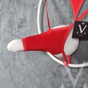 May include: A red thong-style undergarment with a white padded front, displayed on a white circular frame. The garment has thin straps and a tag with a black "V" logo. The background is a textured gray surface.