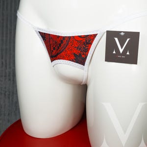 May include: Red and black patterned thong underwear with white trim. The underwear is displayed on a white mannequin. A small tag with a stylized "M" logo hangs from the garment.