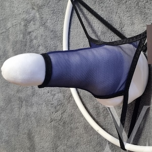 Può includere: Un oggetto a forma di pene in peluche bianco con una copertura in rete blu navy e finiture nere. L'oggetto è sospeso da cinghie nere e da un telaio circolare bianco. Lo sfondo è una superficie grigia strutturata.
