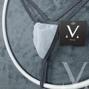 Può includere: Un perizoma in rete bianca con finiture grigio scuro e fascia in vita abbinata. Il capo ha un'etichetta con un logo "V" stilizzato e la scritta "mens jew". Il perizoma è esposto su una superficie grigia.