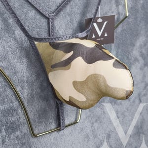 May include: A camouflage-patterned thong with a mesh overlay, displayed on a gold-colored hanger. The thong features a mix of tan, brown, and green hues. A tag with a stylized "V" logo is visible.