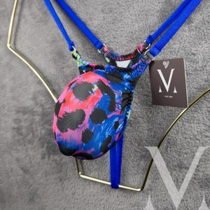 May include: Blue and pink patterned underwear with blue straps. The underwear has a pouch design and is hanging on a gold hanger. The tag on the underwear says "M - one size".