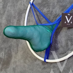 May include: A green mesh jockstrap with blue trim and a white ring. The jockstrap is displayed on a gray fuzzy surface. The brand name "M" is visible on a white tag.