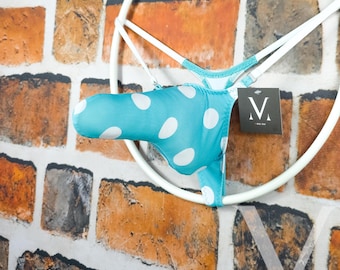 May include: A turquoise and white polka dot thong bikini hangs on a white circular hanger against a brick wall. The bikini has a small tag with the letter "V" on it.