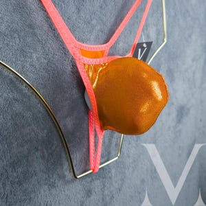 May include: A neon orange and gold bikini bottom with a metallic finish. The bikini is displayed on a gold-colored hanger against a gray background. The bikini has thin straps and a small tag.