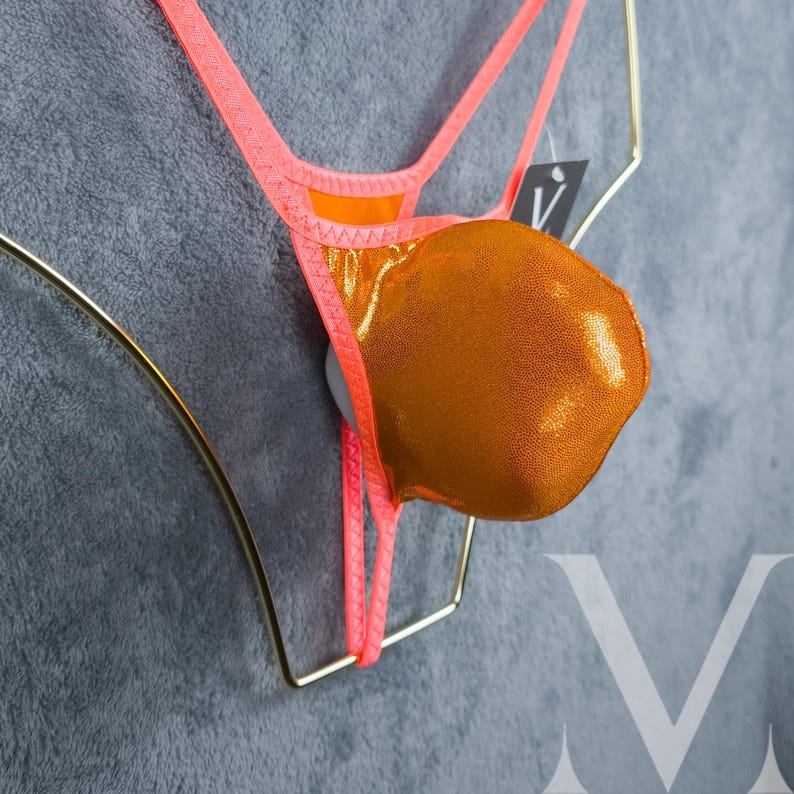 May include: A neon orange and gold bikini bottom with a metallic finish. The bikini is displayed on a gold-colored hanger against a gray background. The bikini has thin straps and a small tag.