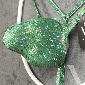 Könnte beinhalten: Ein grünes, paillettenbesetztes Bikini-Oberteil mit dünnen Trägern. Das Oberteil hat eine leuchtende grüne Farbe mit schillernden Pailletten, die das Licht einfangen. Die Träger sind ebenfalls grün und mit Pailletten besetzt. Das Bikini-Oberteil wird auf einer weißen, kreisförmigen Form präsentiert.