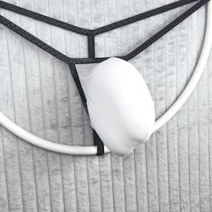May include: A white pouch thong featuring a black elastic waistband and side straps. The garment consists of a solid white fabric front pouch attached to a thin black frame that forms a minimalist structure around the white center section.