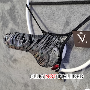 Può includere: Un perizoma nero e argento con un motivo a vortice è esposto su una cornice circolare bianca. Il perizoma ha un piccolo cartellino con un logo. Un tappo rosso a forma di gioiello è attaccato alla cornice. Il testo "PLUG NOT INCLUDED" è visibile.