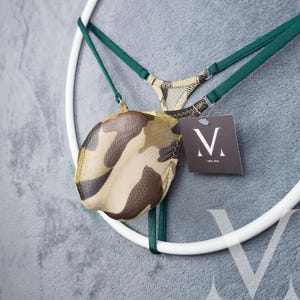 May include: A camouflage-patterned thong with green straps and a tag with the letter "V". The thong is suspended on a white circular frame against a gray background. The camouflage pattern includes shades of brown, tan, and green.