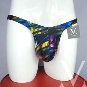May include: A black thong with a colorful, geometric pattern in yellow, green, blue, and purple. The thong is displayed on a white mannequin. A small tag with a white "V" logo is attached.