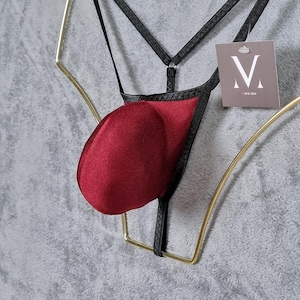 Könnte beinhalten: Ein burgunderroter Satin-Jockstrap mit schwarzen Trägern und schwarzem Besatz. Der Jockstrap hängt an einem goldfarbenen Kleiderbügel. Das Etikett am Jockstrap lautet "M more love".