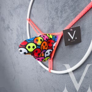 May include: A thong-style undergarment featuring a vibrant pattern of colorful smiley faces. The edges are trimmed with bright coral fabric, and a small tag with a stylized "M" logo is attached.