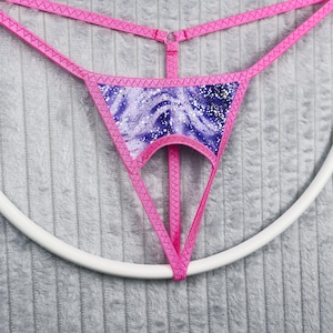 May include: A pink thong with a purple and white patterned front panel. The thong features a delicate design with thin straps and a decorative edge. The garment is displayed on a white circular hanger against a gray textured background.
