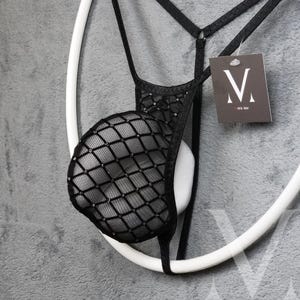 Peut inclure: Lingerie noire avec une pochette en résille à motif losanges et un dos en string. Le vêtement est suspendu à un cintre circulaire blanc. Une étiquette avec un logo "V" stylisé est attachée.