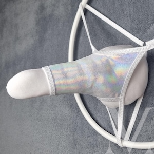 May include: A white and iridescent silver novelty item, featuring a cylindrical shape with a decorative band and a thong-style design. The item is suspended by white straps and a circular frame against a gray background.