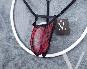 Puede incluir: Un tanga con estampado de leopardo rojo y negro con ribete negro. El tanga está suspendido de una correa negra y de una percha circular blanca. Una etiqueta con un logotipo "V" estilizado cuelga del tanga.