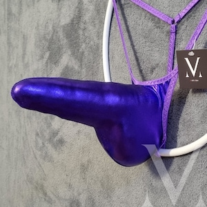 May include: A metallic purple thong-style garment with a phallic shape, suspended from a white ring. The garment has a shiny finish and is attached to a thin, purple strap. A tag with a stylized "V" logo is visible.