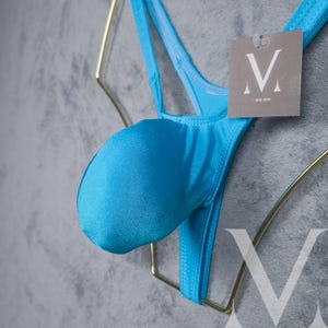 May include: A bright blue thong-style swimsuit with a pouch. The swimsuit is made of a smooth, shiny fabric. A tag with the letter "V" is attached to the garment.