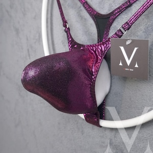 May include: A purple metallic swimsuit with a V-shaped front and a black back. The swimsuit has a shiny, iridescent finish. The brand name "MensKW" is printed on a white tag attached to the swimsuit.