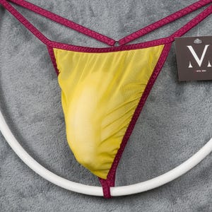 May include: Yellow mesh thong underwear with burgundy trim and straps. The garment is suspended on a white circular hanger, with a tag in the upper right corner. The thong has a V-shape design.