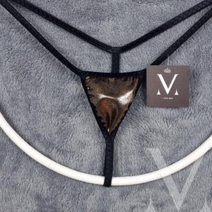 May include: A black and gold thong with a metallic finish. The thong is displayed on a white ring stand. The brand name "M" is visible on a white tag.