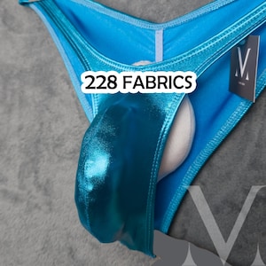 Può includere: Un costume da bagno blu turchese lucido con la scritta bianca "228 FABRICS". Il costume da bagno ha un davanti a forma di V e un inserto imbottito bianco. È attaccata una piccola etichetta con un logo "V".