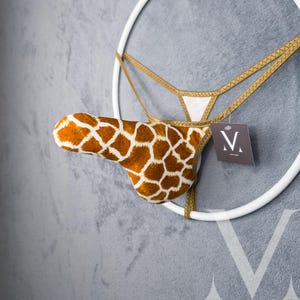 May include: A unique decorative piece featuring a giraffe-patterned thong suspended within a white circular frame. The thong is brown and white, with gold trim. A tag with a logo hangs from the frame, set against a gray textured wall.