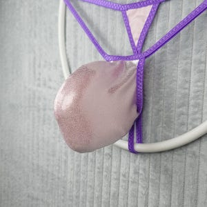 Könnte beinhalten: Ein Tanga mit einer hellrosa, metallisch strukturierten Tasche mit dezentem Punktmuster. Das Kleidungsstück ist mit dünnen, leuchtend violetten elastischen Riemen konstruiert, die eine dreieckige Struktur bilden und einen Kontrast zum Stoff bilden.