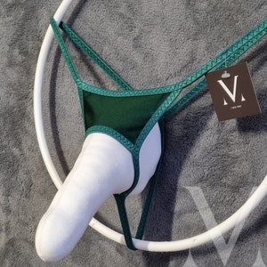 Pode incluir: Um tanga verde escuro com uma frente acolchoada branca e uma alça ajustável branca. O tanga é exibido em um anel branco e tem uma pequena etiqueta com a letra "M".