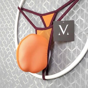 May include: An orange and burgundy thong with a mesh panel and a padded pouch. The garment is suspended on a white circular hanger. A tag with the letter "M" is attached.