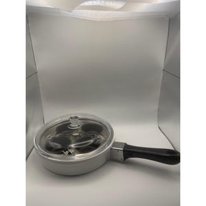 May include: A white round poaching pan with a black handle and a clear glass lid. The interior contains four circular black non-stick poaching cups arranged in a square formation, designed for cooking individual portions of eggs or other food items.