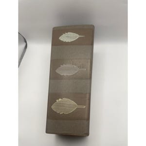 Peut inclure: Un vase rectangulaire en céramique au fini taupe mat, présentant trois bandes horizontales d'une teinte légèrement plus foncée. Chaque bande contient un motif de feuille en relief centré, de ton argenté métallique clair.