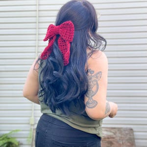 May include: A woman with long dark hair wearing a green tank top and black pants. She has a large red crocheted bow in her hair.