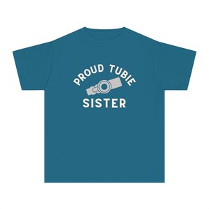 Op de afbeelding: Een tealblauw t-shirt met de tekst "Proud Tubie Sister" in witte letters en een grijze illustratie van een medische buis.