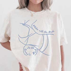 May include: Cream-colored t-shirt with a blue line drawing and text. The design includes the words "Pilates with the Bride" and "Emma's Bach 2026". The shirt is paired with a red and white checkered skirt.