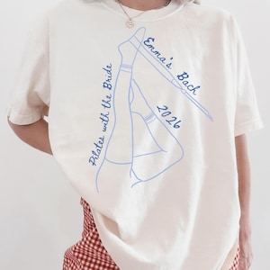 May include: Cream-colored t-shirt with a blue line drawing of legs in socks and the text "Emma's Bach 2026" and "Pilates with the Bride". The shirt is a relaxed fit.